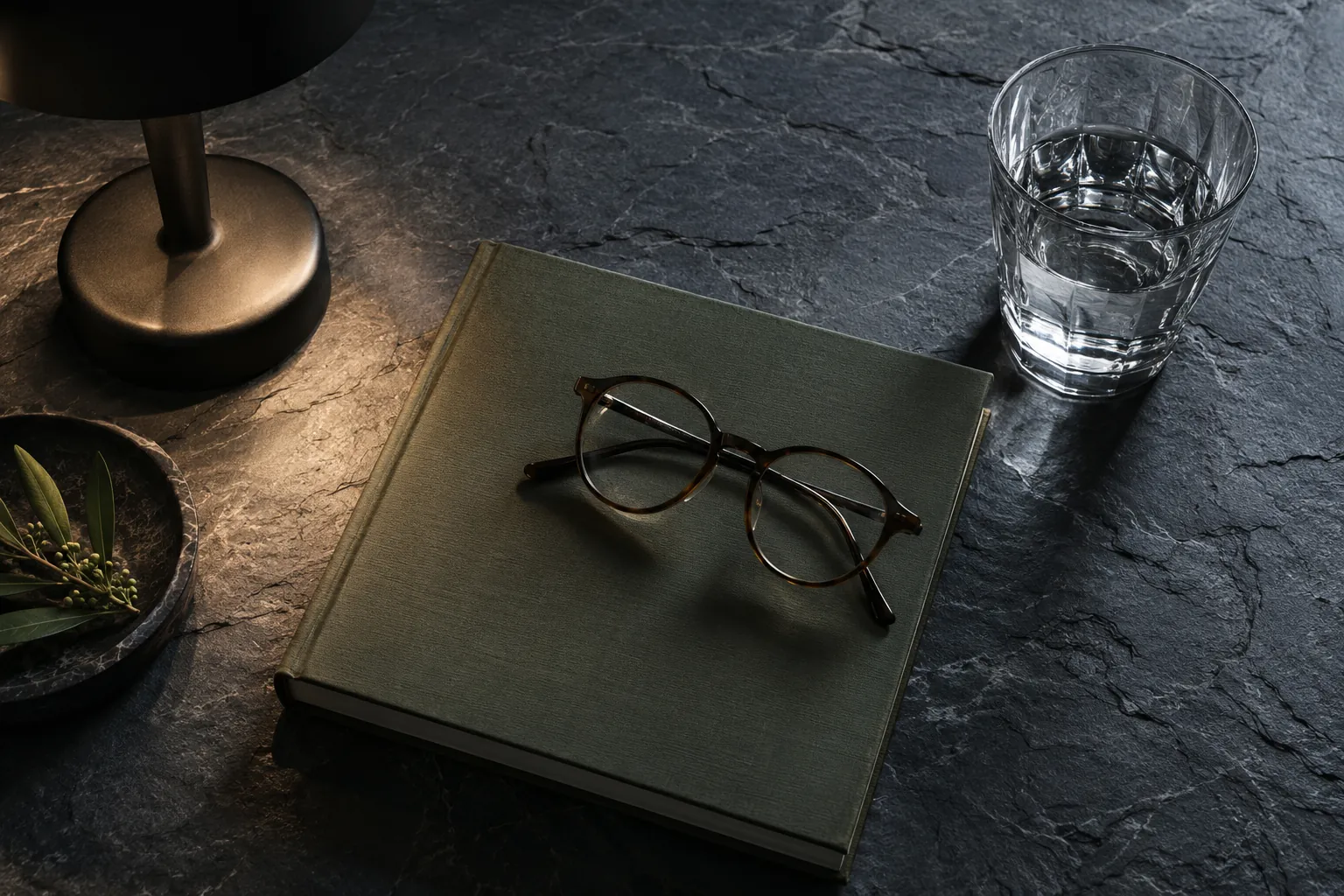 Dark tabletop with a green book, eyeglasses, water glass and part of a black lamp.